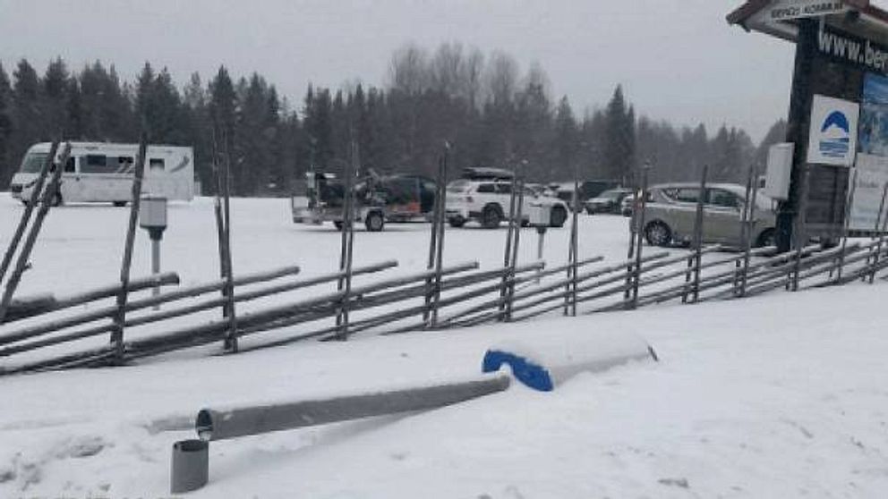 stolpe med fartkamera ligger i snön, kapad. parkering bakom.