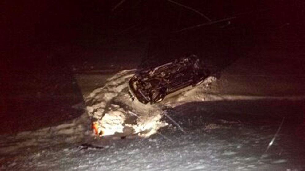 Undersidan av en bil som ligger på en snötäckt is i mörkret. Strålkastarna från bilen lyser upp snön.