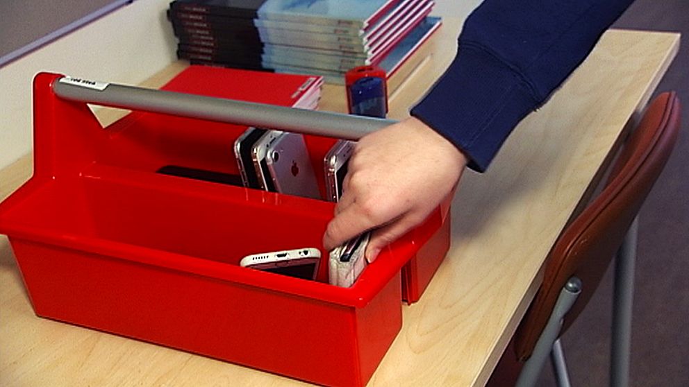 En röd låda står på ett bord. Mobiler står i den, en hand sträcker sig mot en av mobilerna