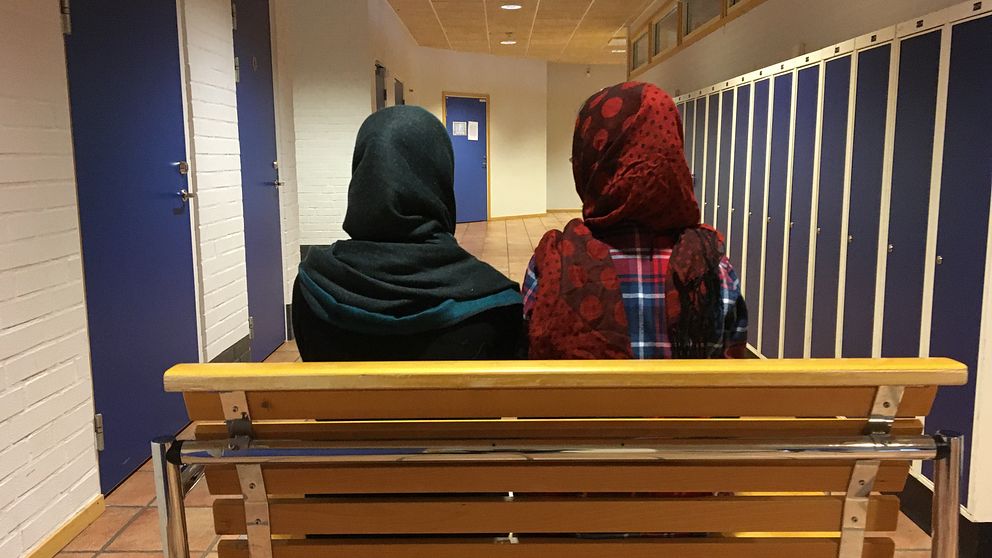 Två tjejer sitter med ryggen emot kameran på en skolbänk och tittar ut mot korridoren.