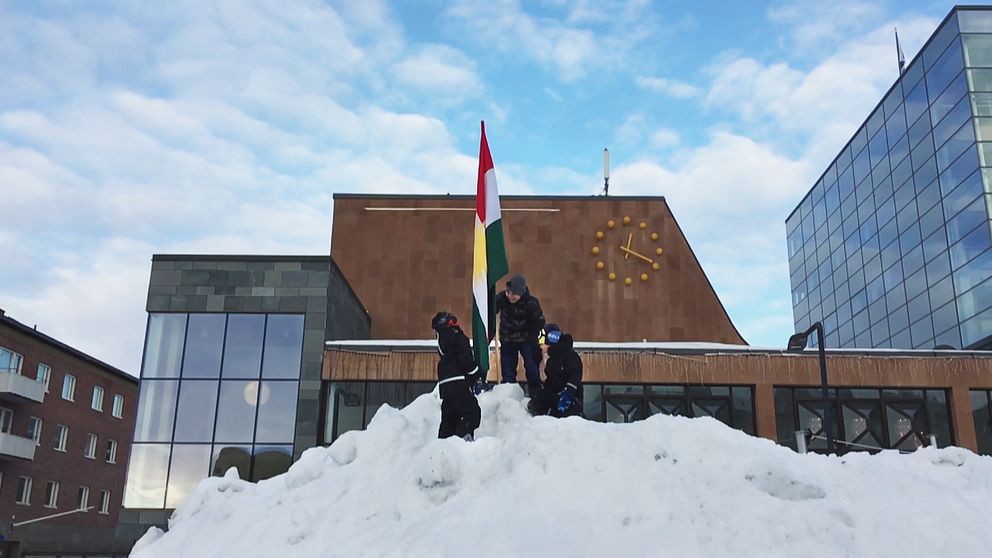 tre barn på snöhög reser flagga, byggnader bakom
