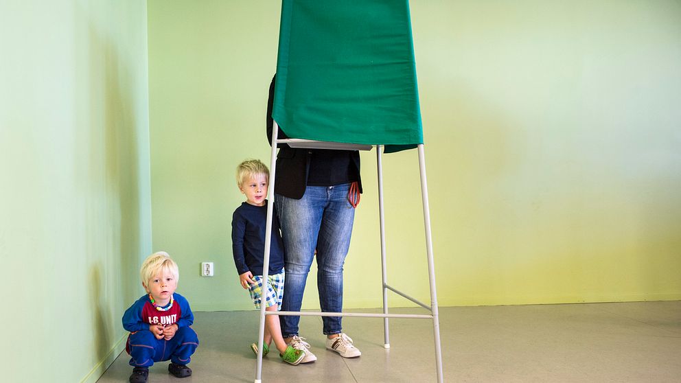 En mamma står i ett röstningsbås medan två barn står bredvid.