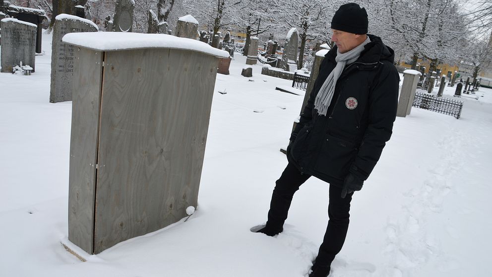 Kyrkogårdschefen Torleif Örn står på en snöig kyrkogård och tittar på en låda av trä som satts utanpå en gravsten. På toppen av lådan har några centimeter snö samlats.