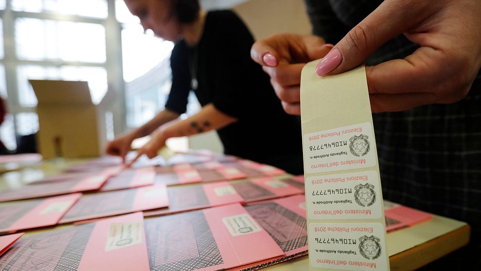 Valarbetande kvinnor står vid ett långt bord och klistrar stämplade lappar på rosa valsedlar inför Italiens val.