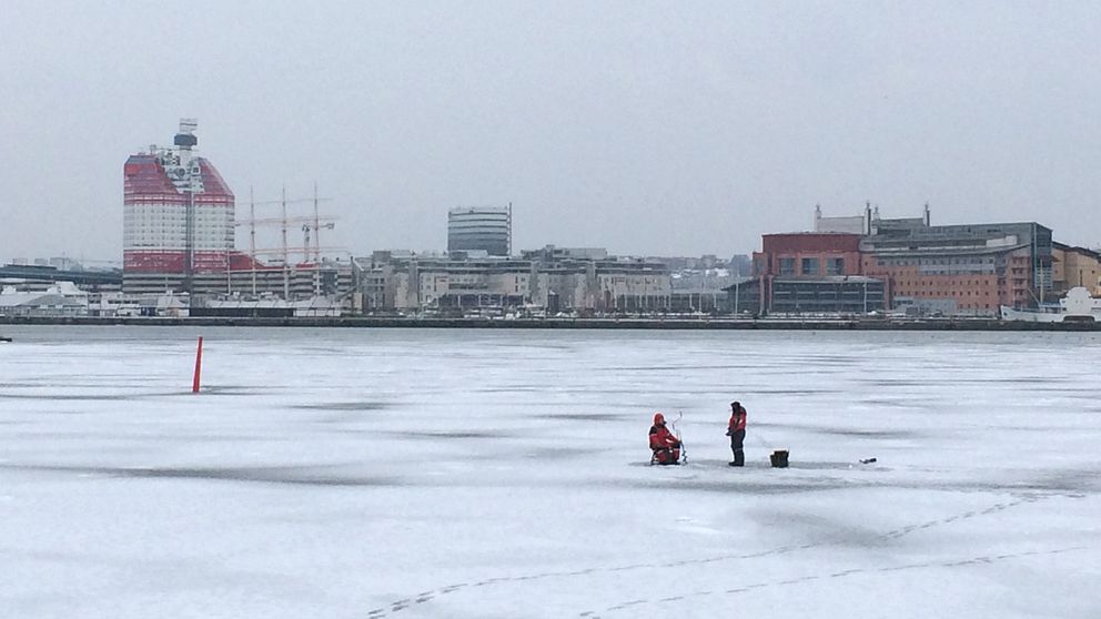 Vintervy, snö och is vid Göteborgs skyline från Hisingen sett, med läppstiftet i horisonten, två personer pimplar ute på isen.