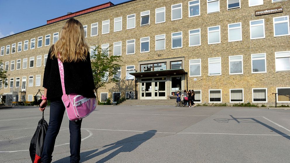 En ensam flicka på väg till skolan över en tom skolgård där en grupp flickor står vid ingången till skolbyggnaden