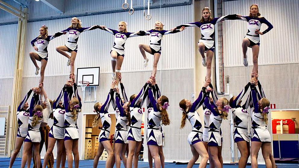En stor grupp cheerleaders stor på golvet och håller upp sex andra som håller i varandras armar och tillsammans bildar en formation
