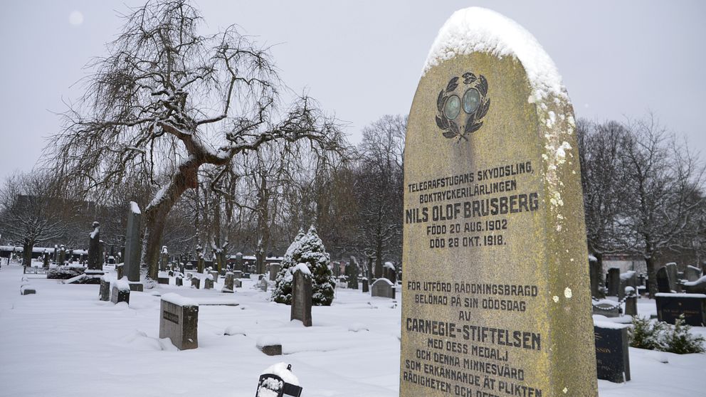 En gravsten med snö på toppen. I bakgrunden syns ett mörkt träd och en kyrkogård. På gravstenen kan man se en medalj, både fram- och baksida, i en inhuggen lagerkrans. På stenen står det: ”Telegrafstugans skyddsling, boktryckerilärlingen Nils Olof Brusberg. Född 20 aug 1902. Död 28 okt 1918.”