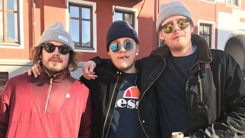 Tre killar med solglasögon, en med en cigarett i munnen och en med en ölburk i handen, står med armarna om varandras axlar.