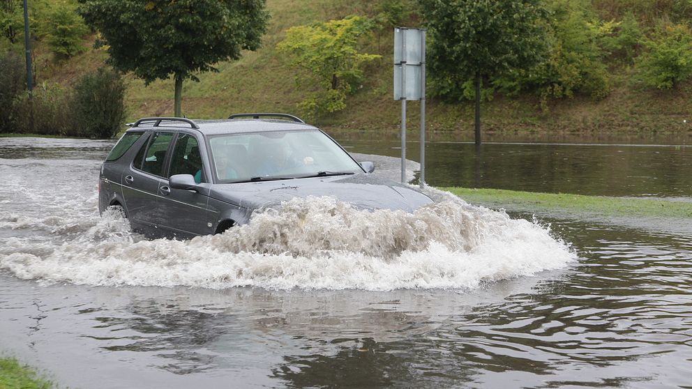 Bil kör igenom översvämmad väg.