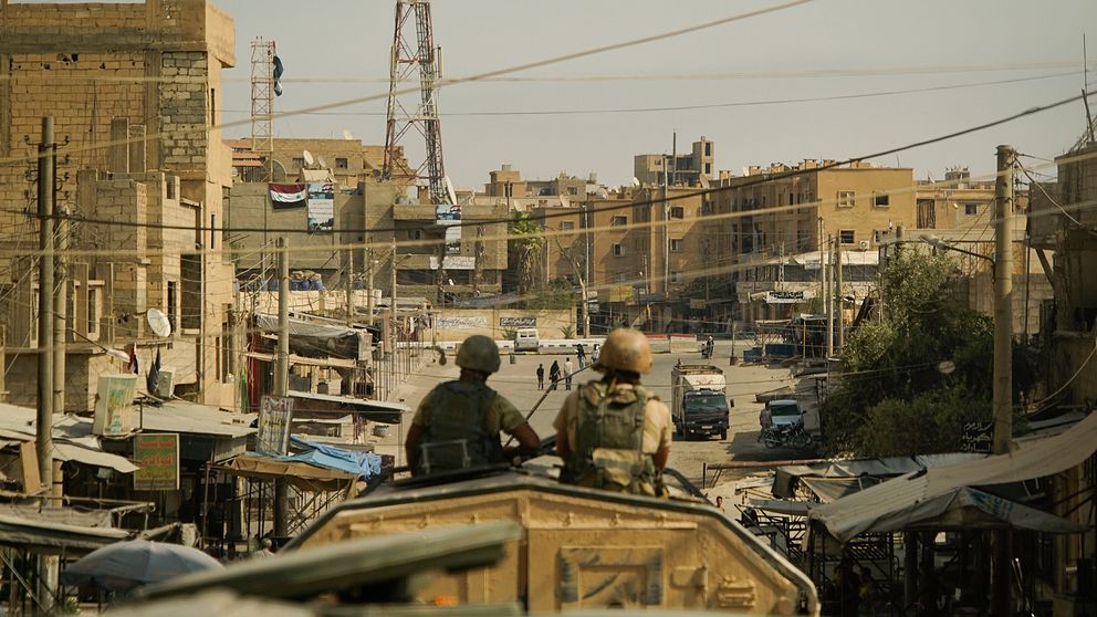 Två män i en stridsvagn blickar ned längs en gata med sandfärgade hus i syriska Deir Ezzor.