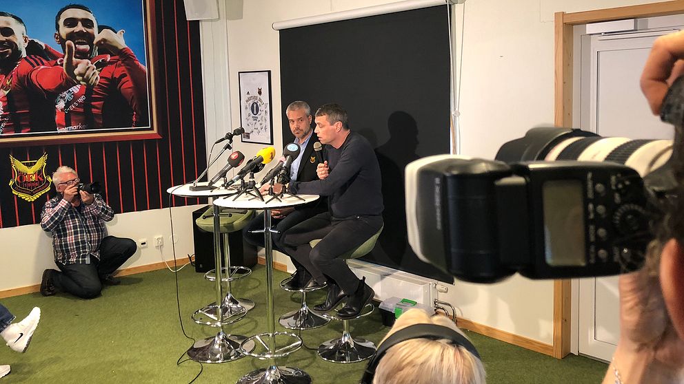 två män sitter vid mikrofoner i ÖFK-lokal, fotografer syns på sidan