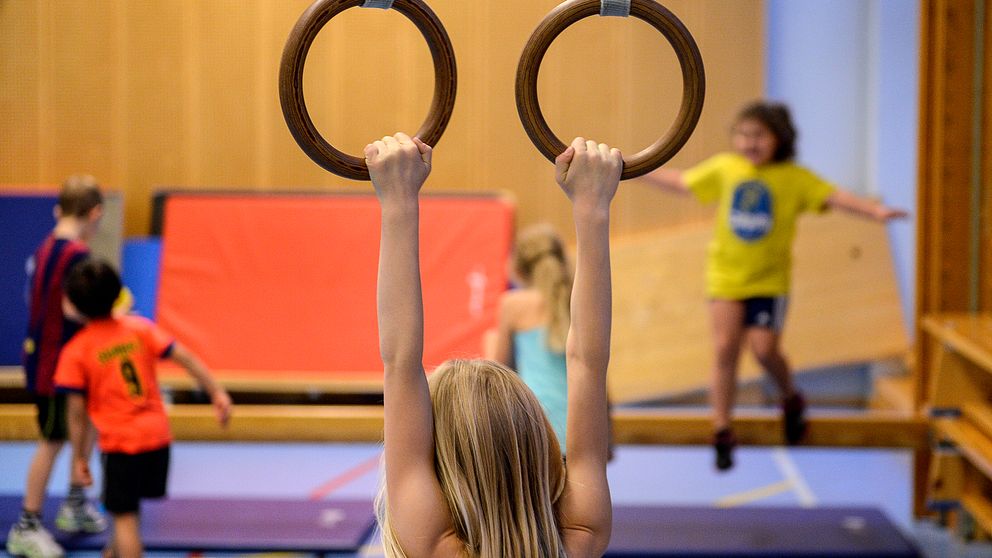 Flicka hänger i romerska ringar i förgrunden, flera barn gymnastiserar i bakgrunden i en idrottshall.