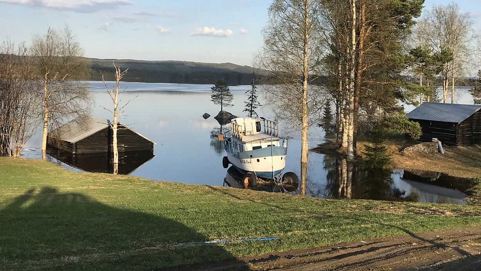 vy över grässlänt ner mot översvämmad sjö, lada i vatten, båt på vagn