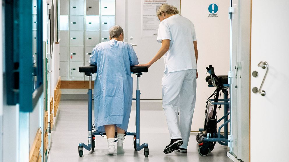 En patient med rullator går i sjukhuskorridoren tillsammans med personal.