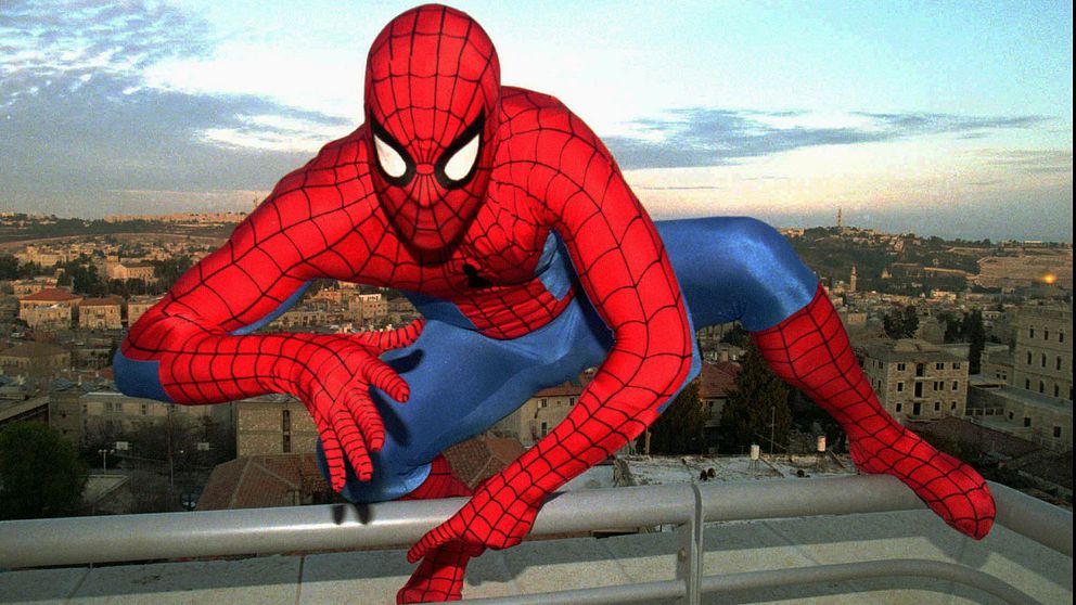 person i Spindelmannen-dräkt klättrar över ett räcke på ett tak, stadsvy bakom