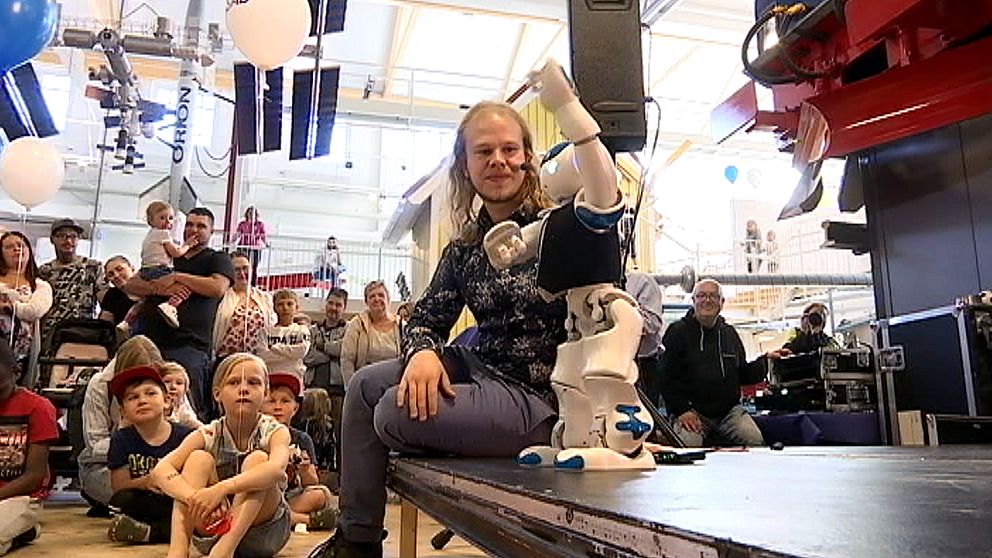 En liten vit robot står på en scen och Fredrik Löfgren, robotutvecklare sitter bredvid.