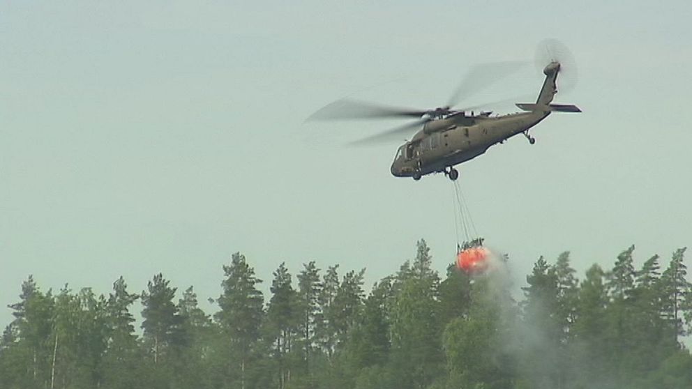 Helikopter flyger över skogen med vattenbehållare under.