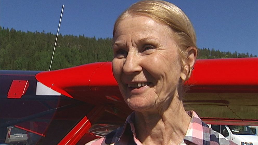 en leende äldre kvinna står ute i solen vid en flygplansvinge