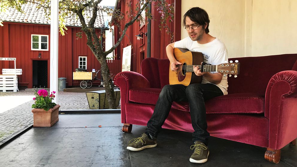 Thomas Jonsson sitter i en röd sammetssoffa. Han håller i en gitarr. I bakgrunden skymtar rödmålade träbyggnader.