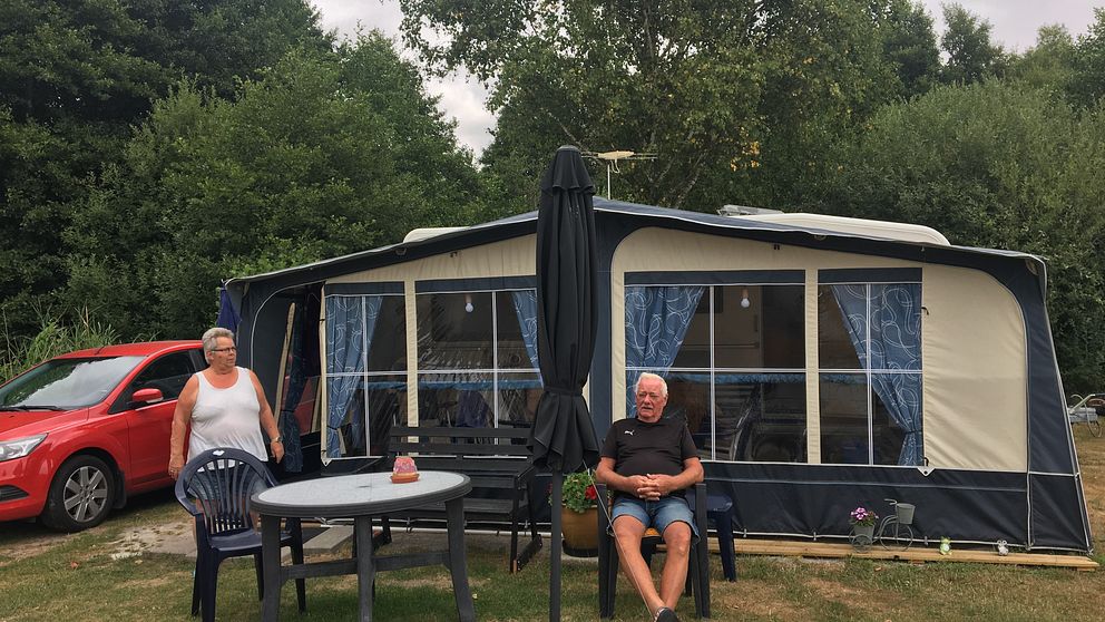 En husvagn med förtält uppsatt på en camping. En man sitter i en solstol framför och en kvinna går ut ur förtältet.