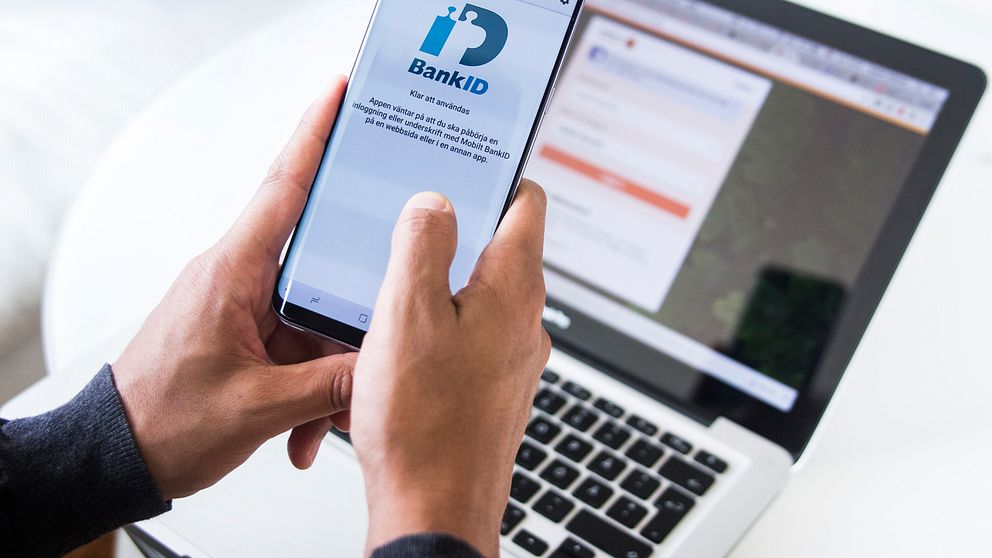 Två händer håller i en mobil framför en dator. På mobilskärmen syns mobilt bank-id:s logga.