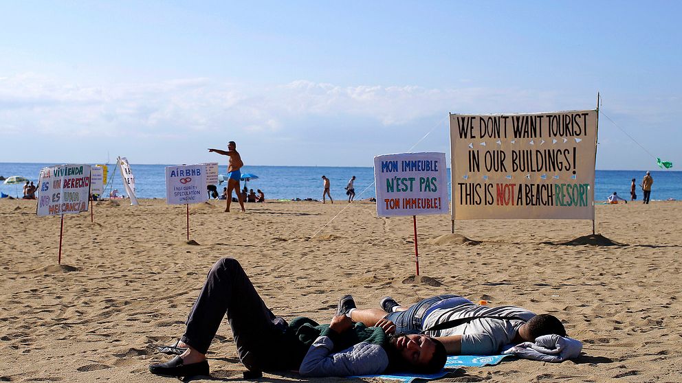 Två män ligger på en spansk sandstrand omgivna av skyltar som protesterar mot att turister bor i vanliga bostadshus.