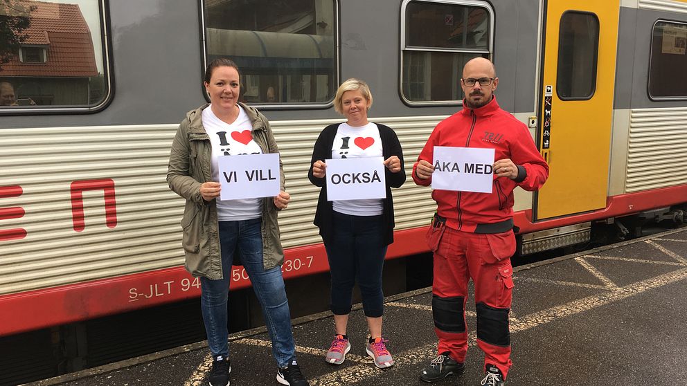 tre protesterande med A4-papper med texten ”VI VILL OCKSÅ ÅKA MED” framför ett Krösatåg.
