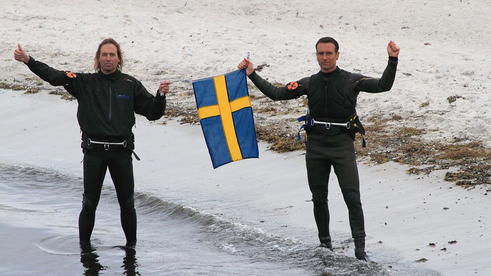 De två vindsurfande svenskarna står med en svenska flagga mellan sig.