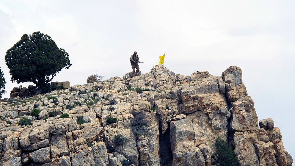 En hizbollahsoldat står på ett berg i Syrien, bredvid honom vajar organisationens flagga.