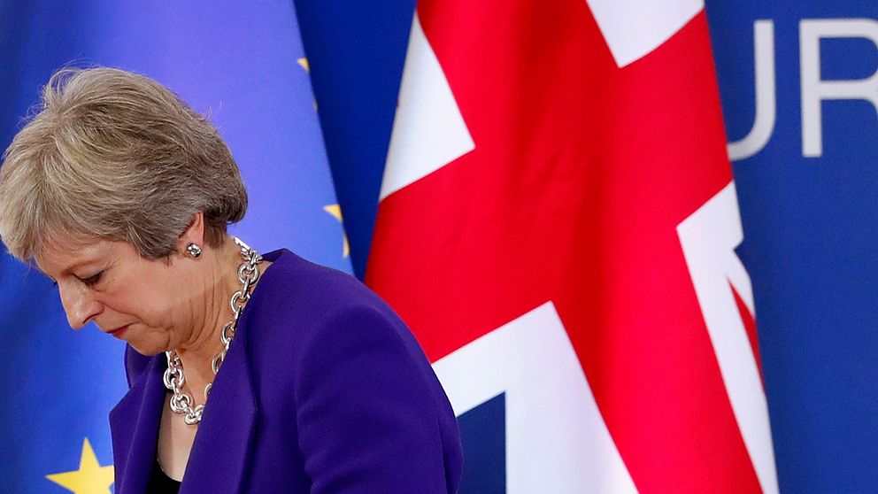 Theresa May tittar ner. I bakgrunden syns Storbritanniens och EU:s flaggor.