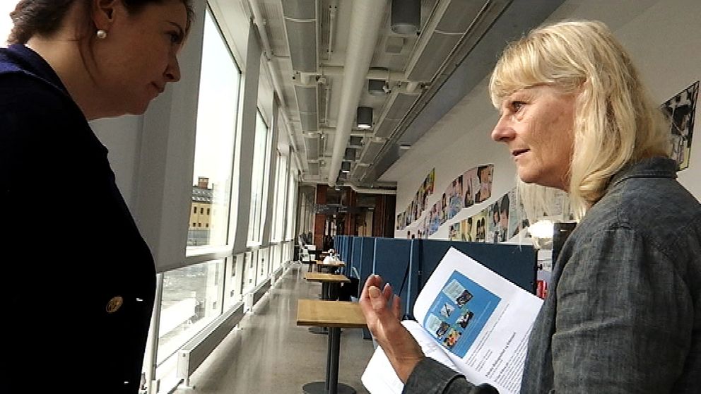 Två personer står och diskuterar, med papper i handen. I bakgrunden en korridor på högskolan.