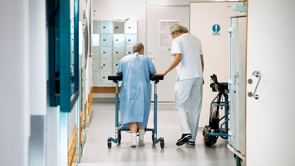 En kvinnlig patient med en rollator får hjälp av personal att ta sig fram i en korridor på ett sjukhus