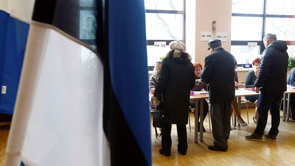 En kvinna och två män registrerar sig för att rösta i Estlands parlamentsval. Estlands flagga i förgrunden.