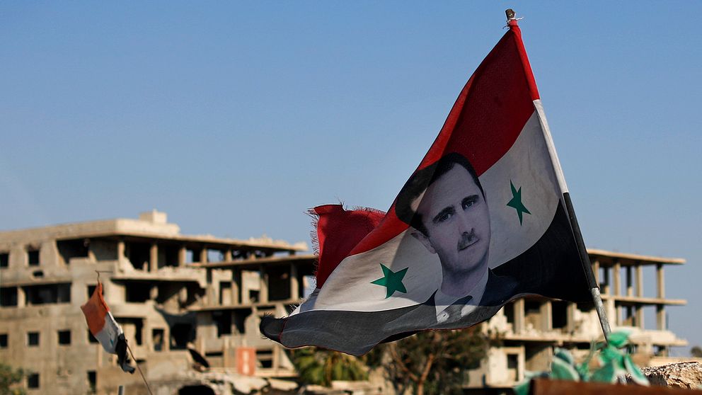 En syrisk flagg med bild på president Bashar al-Assad, i bakgrunden sönderbombade byggnader.