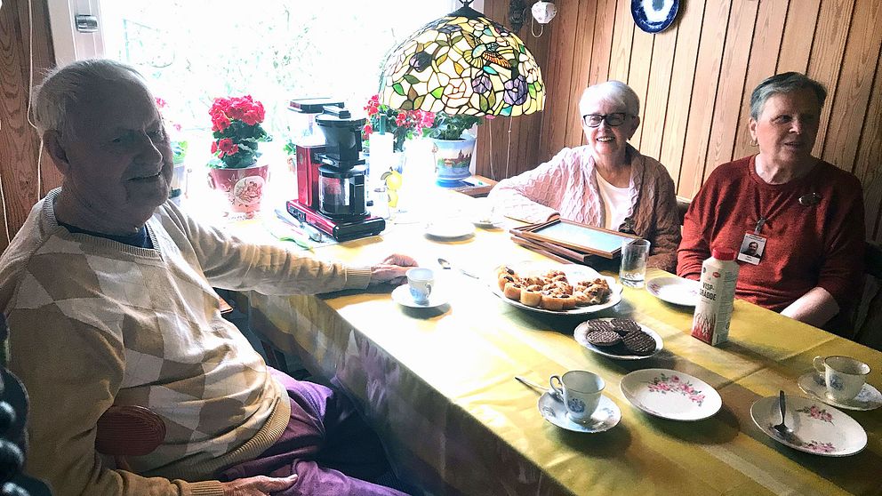 En äldre man och två äldre kvinnor sitter tillsammans vid ett fika bord.