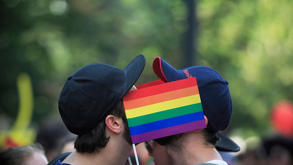 Två män kysser varandra bakom en regnbågsflagga.