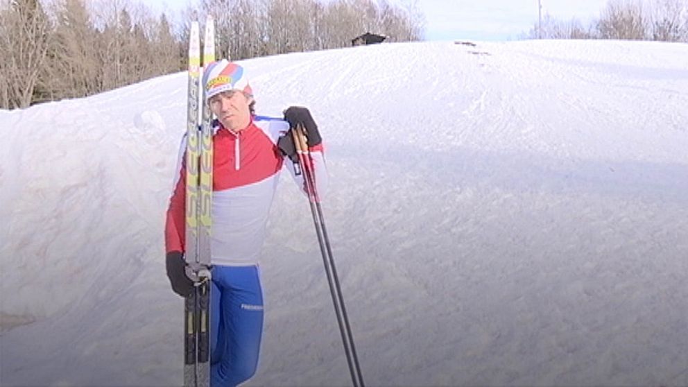 Man som kisar av solen, har randig tight mössa och tighta blå/röd/vita träningskläder. Ett par skivstavar i högerhanden, med skidor lutandes mot axeln. En snötäckt kulle i bakgrunden.