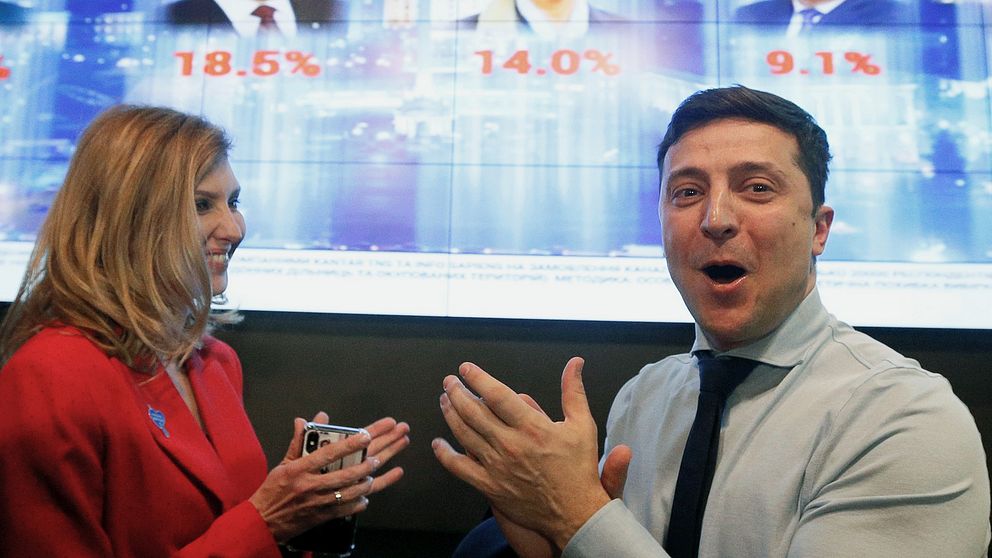 Bild på komikern och presidentutmanaren Volodymyr Zelenskyj och hans hustru framför en skärm som visar valresultaten för de olika kandidaterna.