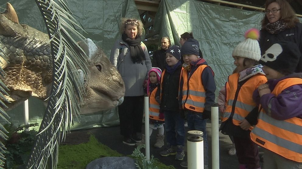 Flertal barn med två lärare och en dinosaurie.