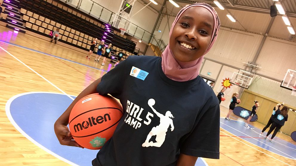 Ikran Said håller en basketboll under armen och ler mot kameran. Hon har på sig en t-shit där det står ”girlscamp” och en rosa slöja. Bakom henne står flera tjejer och övar.