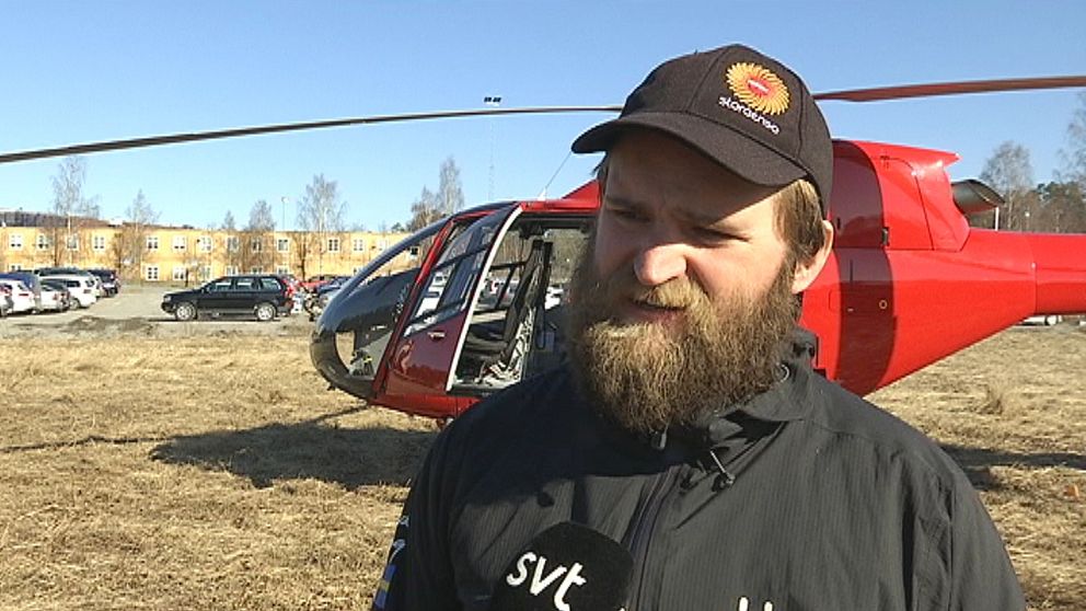 en man i keps och jacka står framför en helikopter på gräsyta, bebyggelse i bakgrunden