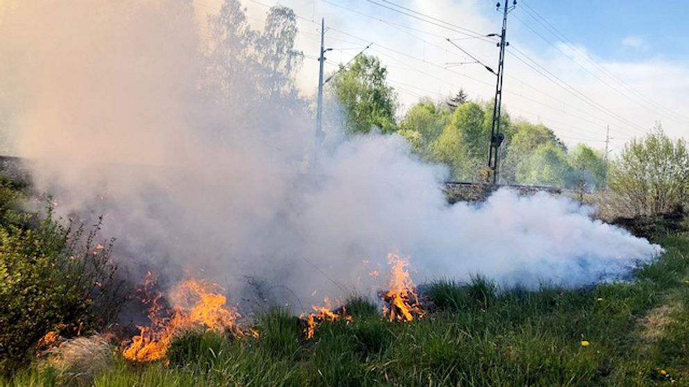 Brand som sprider sig i gräset på en banvall. Grå, tjock rök sprider sig från elden.