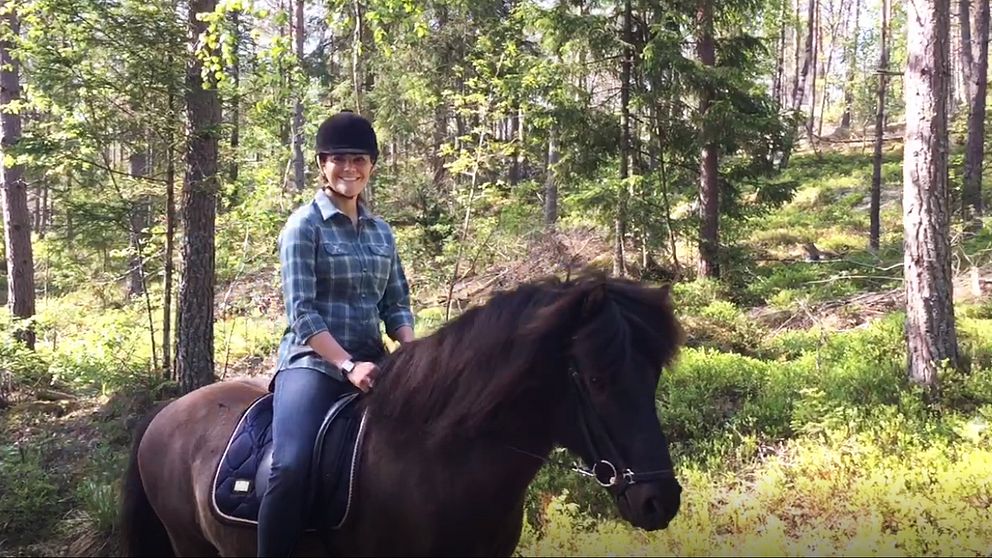 Kronprinsessan Victoria rider in i Sörmland på ryggen på en mörkbrun islandshäst. Hon ser glad ut, är iklädd rutig skjorta och ridhjälm.