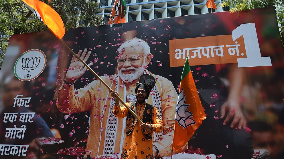 En man viftar glatt med en orange flagga framför en stor bild på indiske premiärministern Narendra Modi.