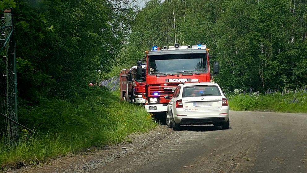 Två brandbilar syns på en grusväg med lövskog på båda sidor. En vit bil står parkerad framför räddningsfordonen.