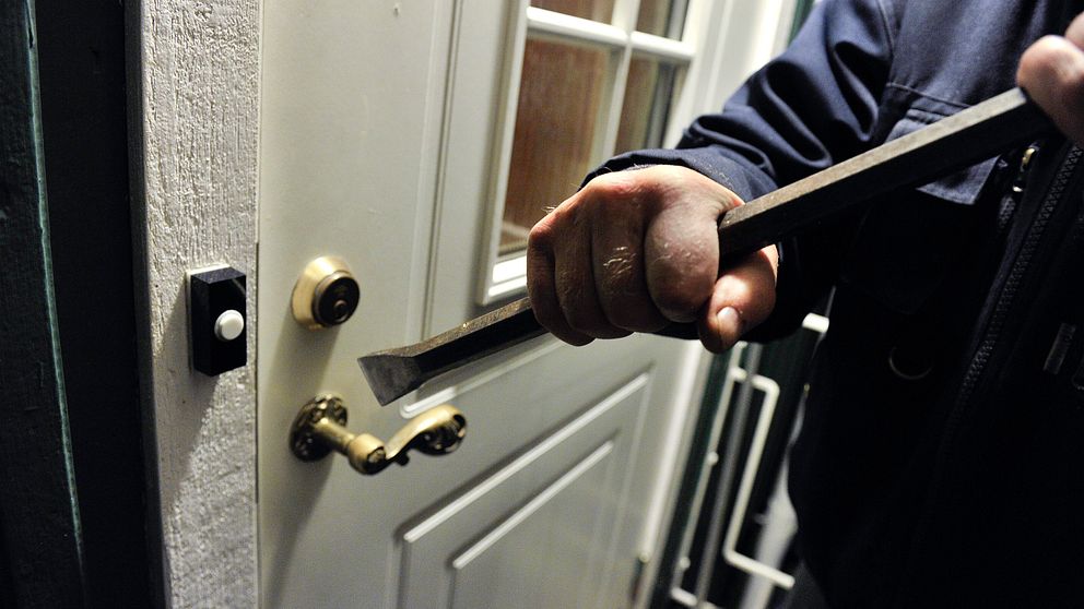 En man står vid dörren till en villa med en kofot i händerna, på väg att göra inbrott.