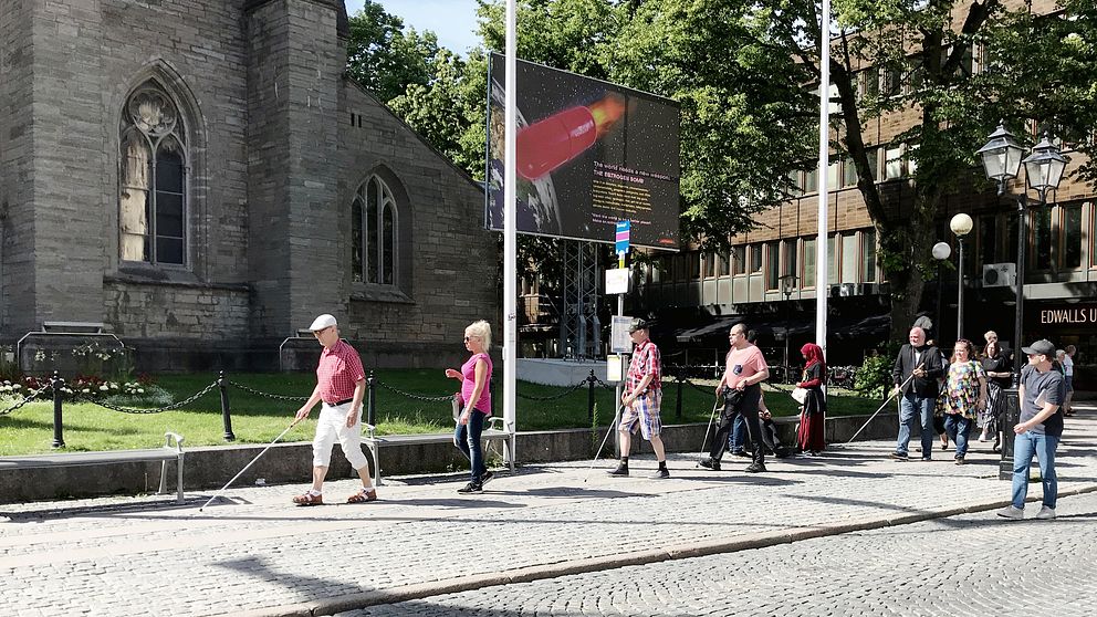 Flera ur Synskadades rikförbund passerar Nicolaikyrkan på Stortoget. De går med käpp, och på rad.
