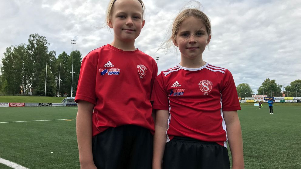 Två flickor står med fotbollsplan i bakgrunden