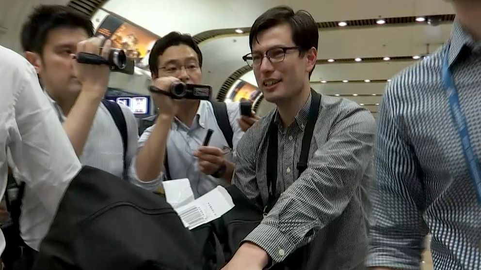 29-årige Alek Sigley anländer till flygplatsen i Peking i Kina omringad av journalister. Bilden är tagen ur en video.